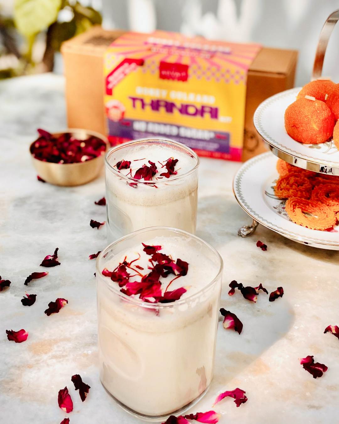 2 glass tumblers of Thandai placed on a marble table with some sweets in the background.